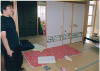 和室の障子が破れ畳の上に布団やまくら、リモコンなどが散乱し男性が立っている写真