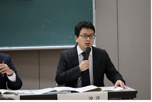 黒板の前の席に座り右手にマイクを持ち前方を見ながら話をしている金城氏の写真