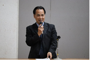 演台に立ち右手でマイクを持って話をしている前泊氏の写真