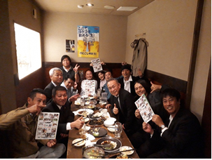 居酒屋さんで食事をしながらパンフレットを持ってピースサインをしている12名の皆さんの写真