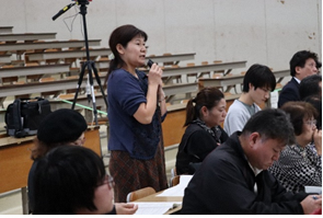 一番後ろの席に座っていた報告会に参加した女性が立ってマイクを両手で持ち意見を述べている写真