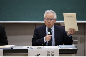 黒板の前の席に座り右手にマイク、左手に資料を持ち話をしている名嘉座氏の写真