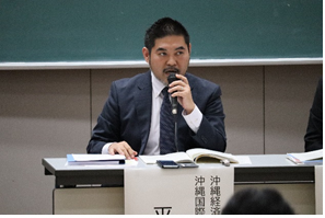 黒板の前の席に座り右手にマイクを持ち、左手を机の上に置いて前方を見ながら話をしている平敷氏の写真