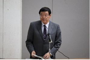 演台に立ち資料を持ちマイクの前で話をしている前津学長の写真