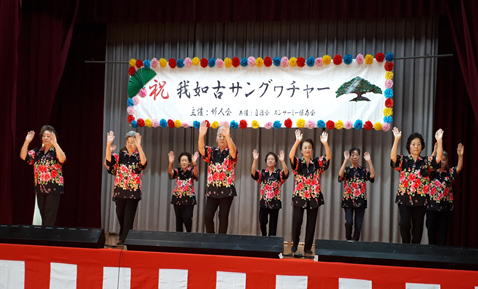 黒のアロハシャツを着た老人クラブの方々が、手のひらをみせながら両手を上げて踊っている写真
