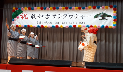 太鼓とバチをもち踊っている4人の女性、日の丸が描かれた扇子を片手に踊っている数名の女性が向かい合っている写真
