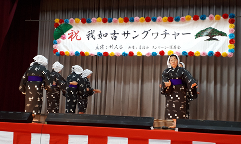 舞台の前方をむいている人達と後ろ姿の、黒に白地で格子模様が描かれたお揃いの着物姿の婦人達が両手を下におき踊っている写真