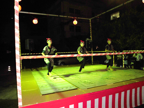 舞台上で3人の女性が赤のねじり鉢巻きをして右足をあげて踊っている写真
