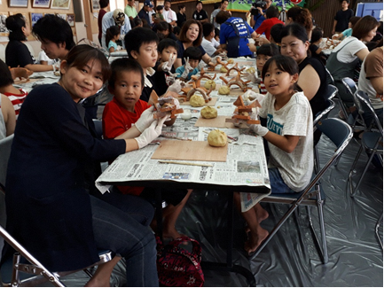 新聞紙の上に置かれた板に材料となる粘土が丸められて準備されている机にグループごとに座り、参加者の人達が笑顔でカメラをみている写真