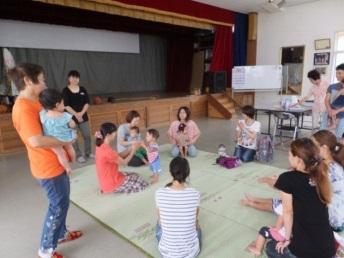 体育館内の敷物の上に親子数組が座り、その周りに数名の人達が立ちながら子供を抱いたり見守っている写真