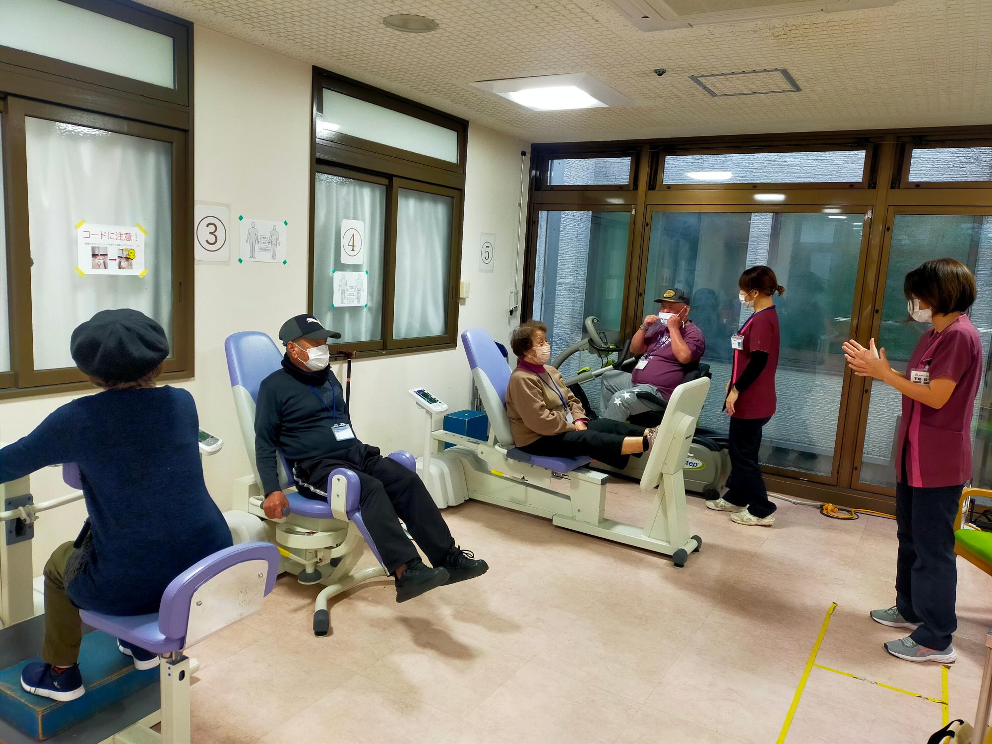 おもと園パワーリハ教室。4名の参加者はトレーニングマシーンを用いて運動中。指導者二人はリズミカルに回数を数えている。