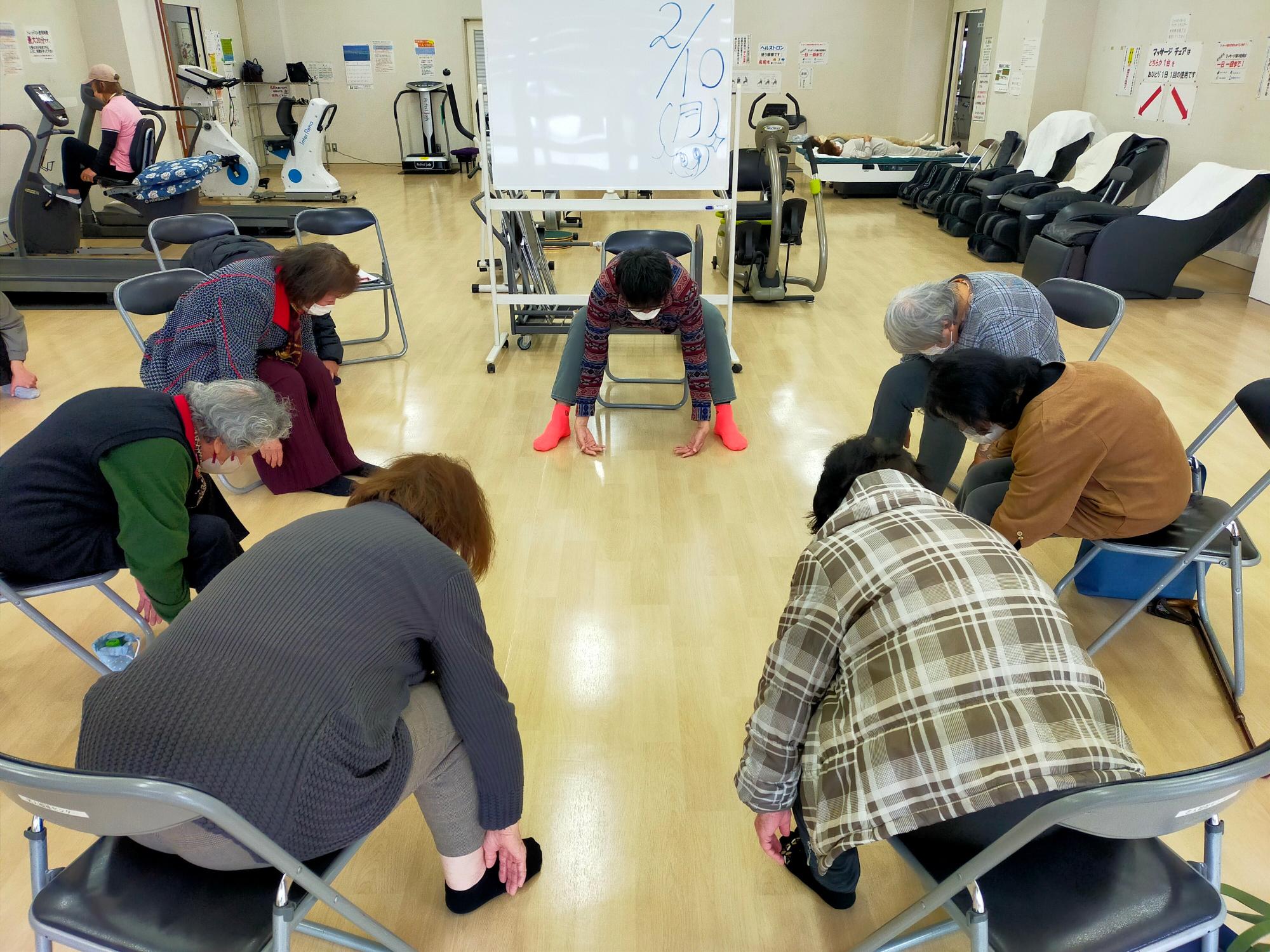 疼痛改善教室。指導者と参加者は椅子に座り、円になっている。股関節を使って前に倒れる「前まげ」の動作をしている。