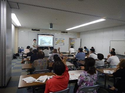 教室の前でスクリーンを使い説明している男性が立っており、横2列に並べられた長机に参加者が座っており、男性の方を向きながら話を聞いていたり、手をあげている参加者の方々が写っている写真