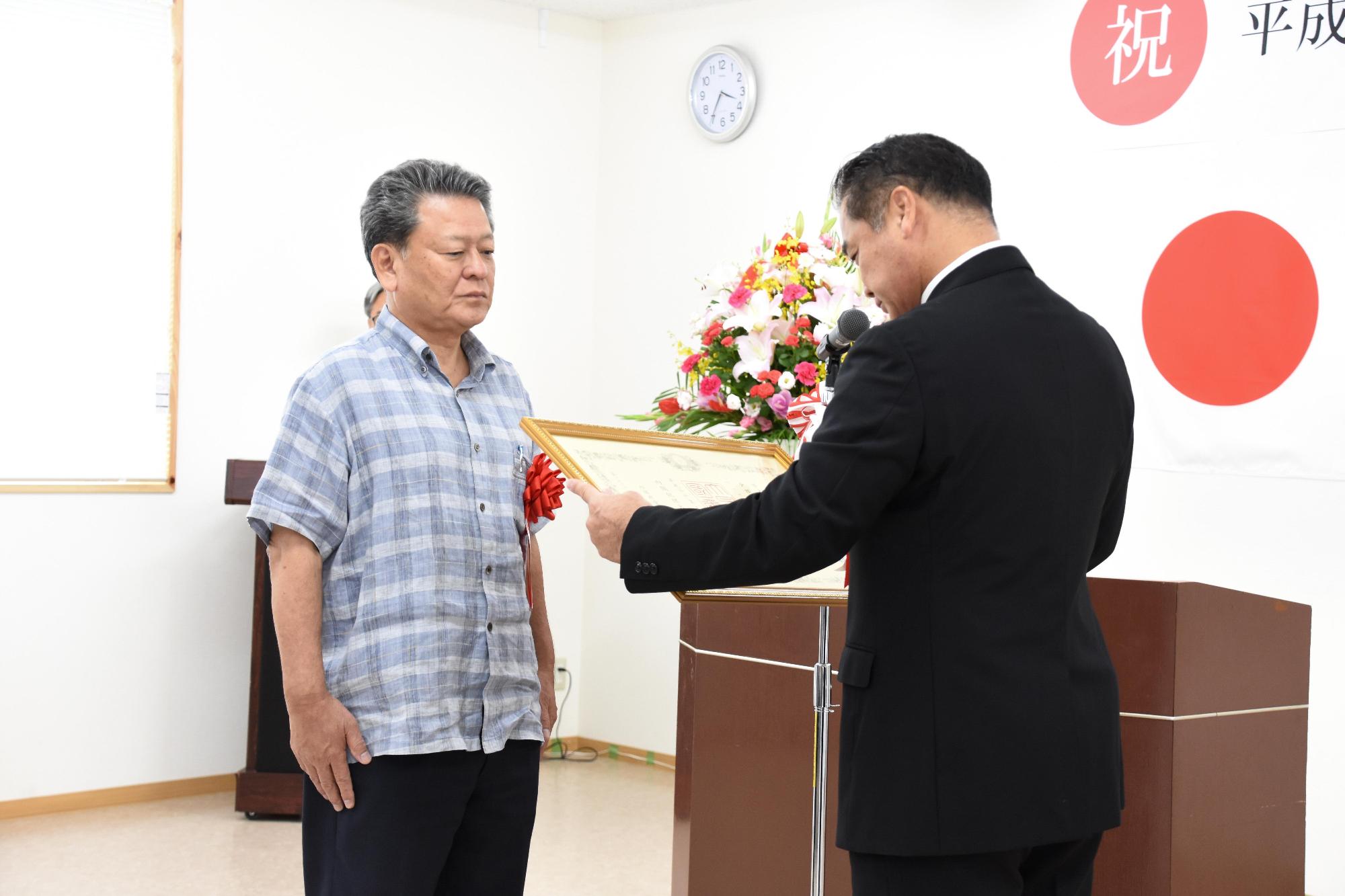 額縁に入った表彰状を読んでいる佐喜眞市長と表彰を受けている男性の写真