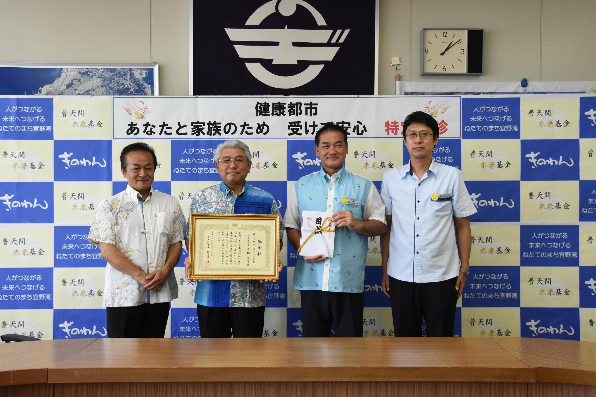 額縁に入った賞状を両手で持っている石松社長と目録を持っている佐喜眞市長と両隣に立っている関係者の方々の集合写真