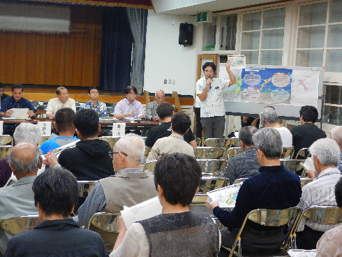 参加者の方々と関係者の方々が向かい合って椅子に座っており、うち関係者の1名の方が白板に貼られた資料を指さしながら説明している写真