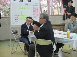 マイクを左手に持ち席に着いている男性が右手で指を指しながら話をしている写真