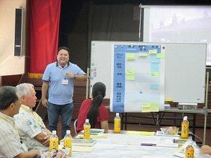 男性が前方に置かれたホワイトボードの横に立ち、席に座っている参加者たちの方を向いて話をしている写真