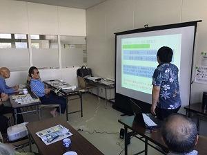 アロハシャツを着た男性がスクリーン画面を使って説明していているのを聞いている参加者の写真