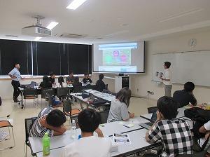 プロモーションビデオをスクリーンに映し説明している男性と、参加されている大学生の写真