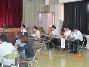 椅子に座っている女性がマイクで話をしていて、その後ろにワイシャツの男性3人が座っている活動報告の写真