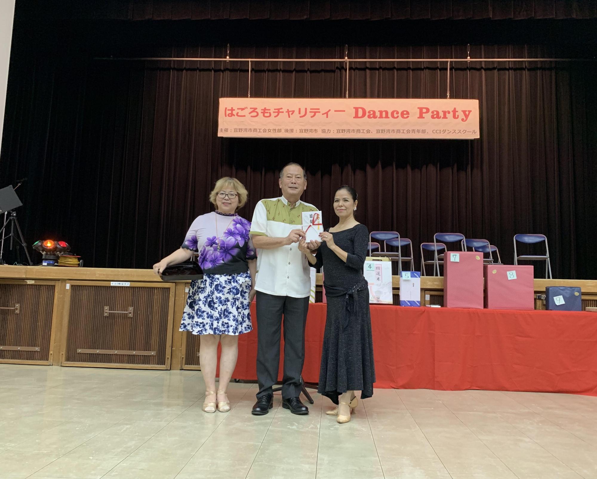 松川市長と黒いドレスを着た女性が2人で目録を持っており、松川市長の左隣には眼鏡をかけ、花模様の洋服を着た女性が写っている写真