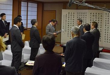 7名ほどの関係者がいる室内で文書を読み上げている市長の写真