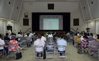 前方にスクリーンがあり半分ほどの席に市民が座っている会場内の写真