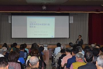 舞台上のスクリーンには資料が映され舞台下で多くの市民が報告を聞いている写真