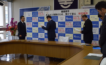会議室で5名の関係者が起立し向かい合って文書を読み上げている写真