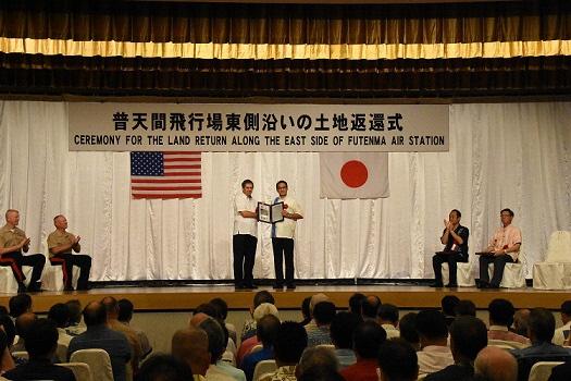 外国の男性と日本の男性が返還の証明文書を持って並び拍手がおこっている写真