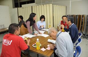 立ち上がり、テーブルの上の模造紙に書き込もうとしている1人の女性と、同じテーブルで話し合いをしている4人の男性と1人の女性の写真