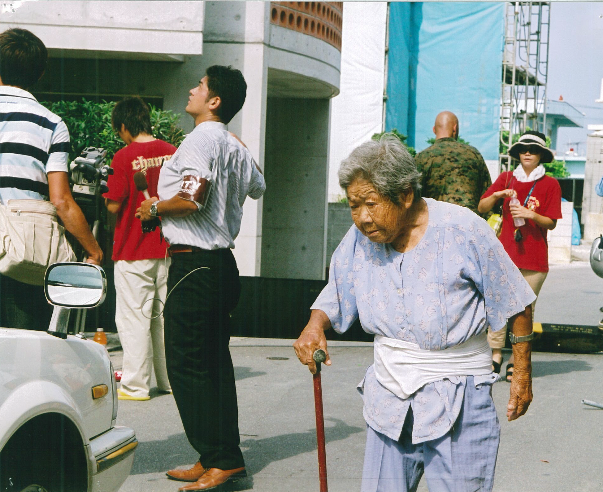 マイクをもった男性の後ろを紫のパジャマ姿の年配の女性が杖をついて歩いている写真