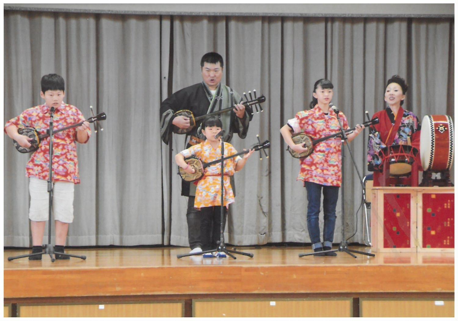 三線の演奏をしている3人の子供、1人の男性、太鼓をたたいている1人の女性の写真