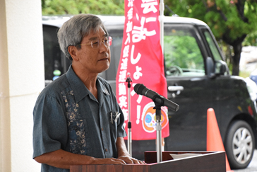 グレー色のシャツを着た社会福祉協議会長が市役所玄関前に設置された演壇の上に両手を置いてマイクの前で話しをしている写真