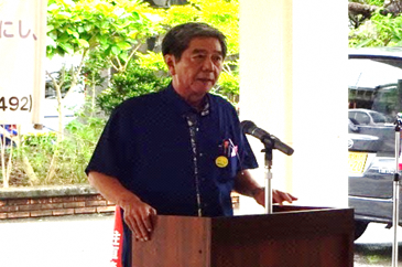 青色のシャツを着た中原区の森田自治会長が、市役所玄関前に設置された演壇の端に手を添えてマイクの前で話しをしている写真