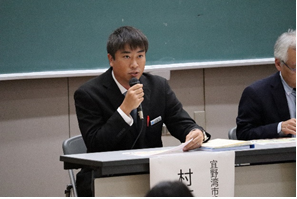黒板の前の席に座り右手にマイク、左手で資料をを持って話をしている村木氏の写真