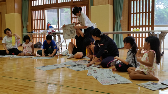 体育館内に新聞紙を広げ子供達が中央にいる人からスリッパのつくり方を聞いている写真
