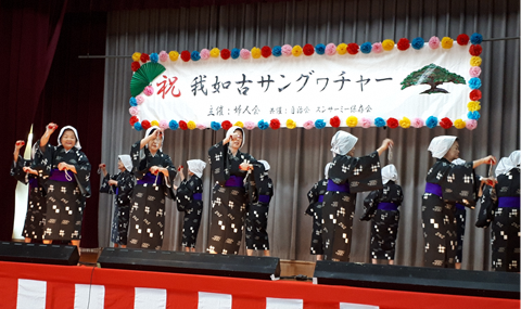 舞台上で黒に白地で格子模様が描かれたお揃いの着物姿の婦人方が右手を上に左手を腰あたりにおいて踊っている写真