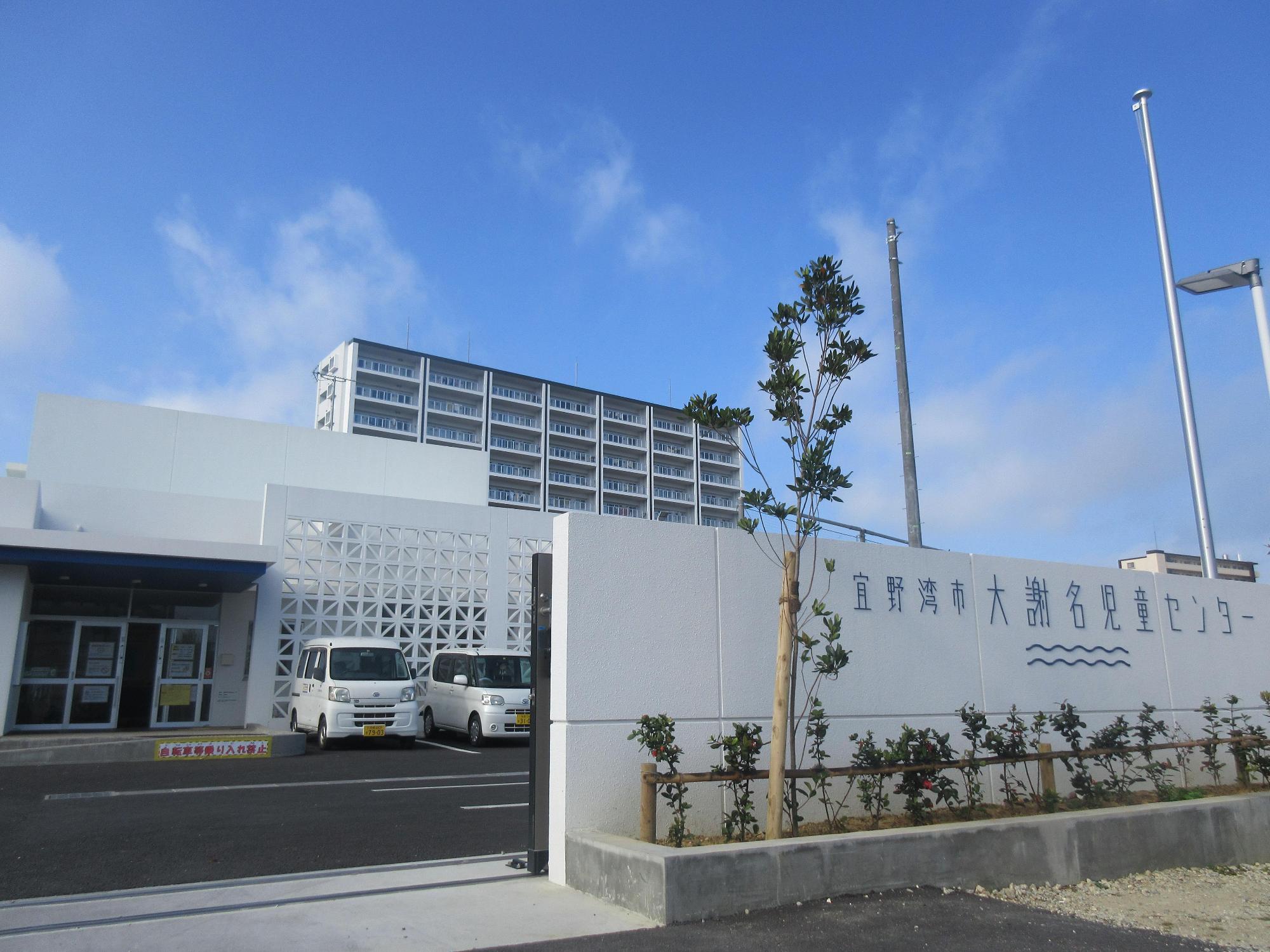 敷地入り口前に宜野湾市大謝名児童センターと書かれてある白い壁と植栽があり、壁の奥に白い外壁の児童センターが写された写真