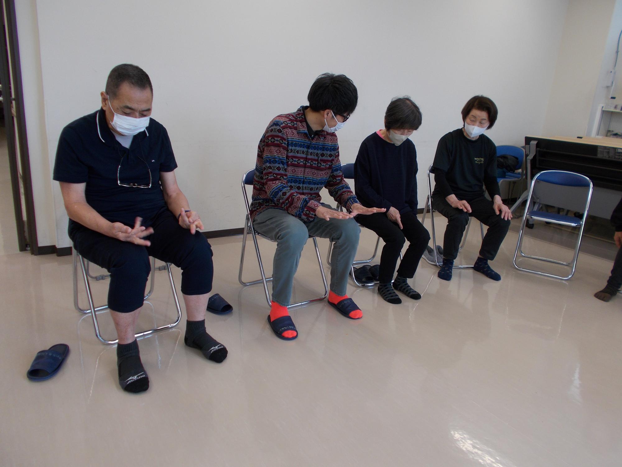 がんじゅう広場。指導者と参加者数名が椅子に座っている。指導者は動作法を手を用いて指導している。