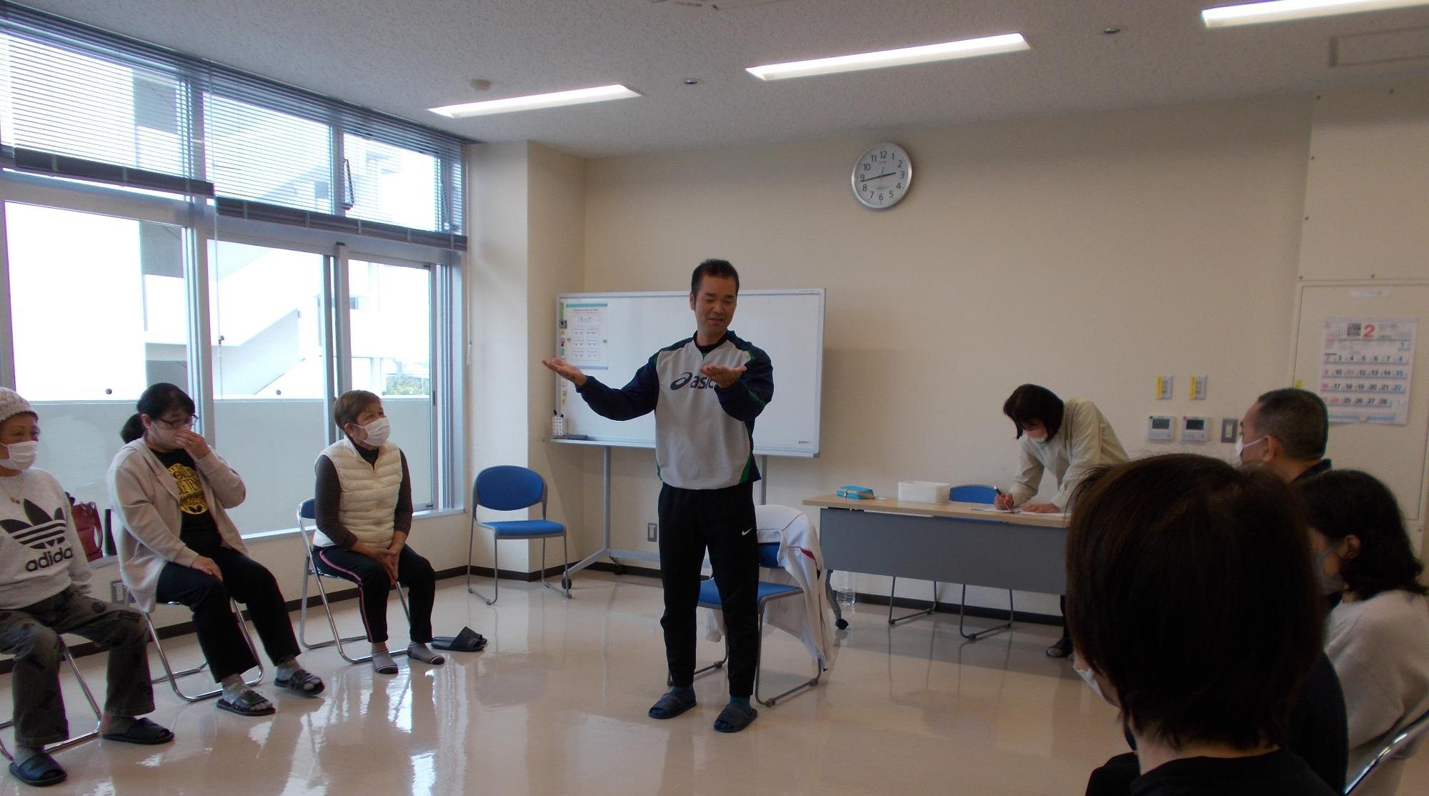 がんじゅう広場。指導者は立ちながら動作法について説明している。参加者は椅子に座り、お話を聞いている。