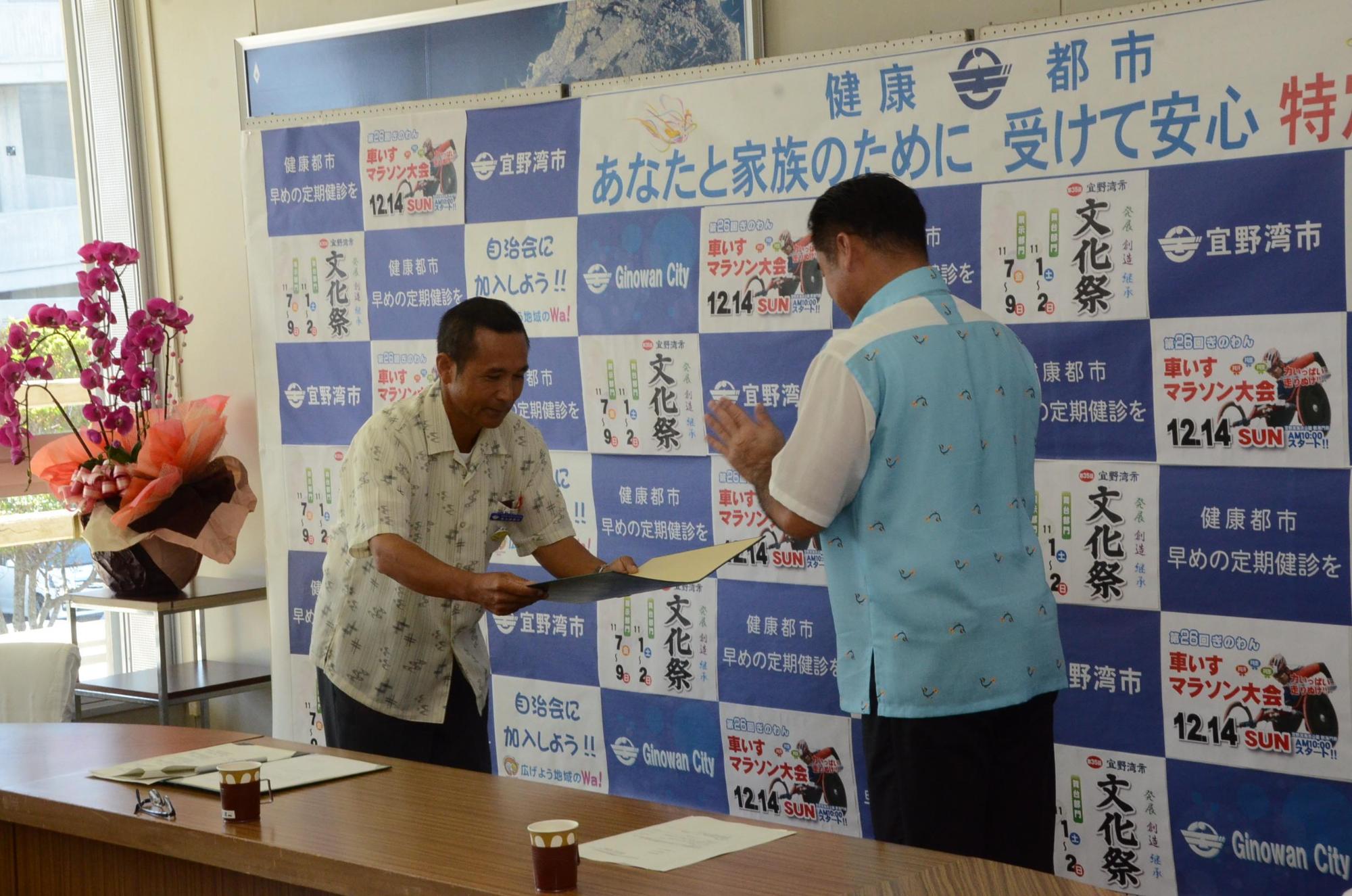 認定証受書式にて宇地泊区自治会 富名腰 義政会長が市長より賞状と受け取っている写真