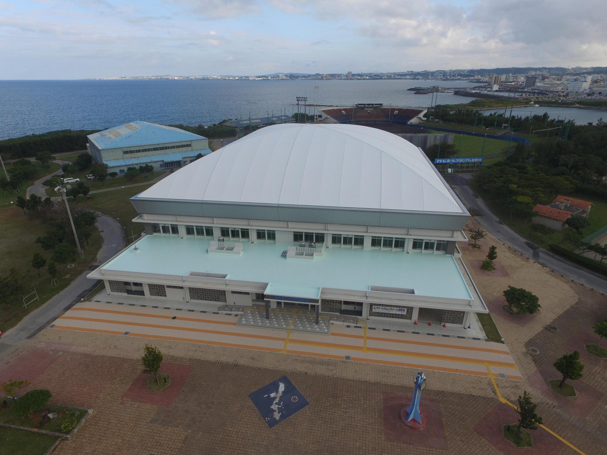 海の近くにある屋根が台形の形をした「宜野湾市立多目的運動場」を上空から撮影した写真