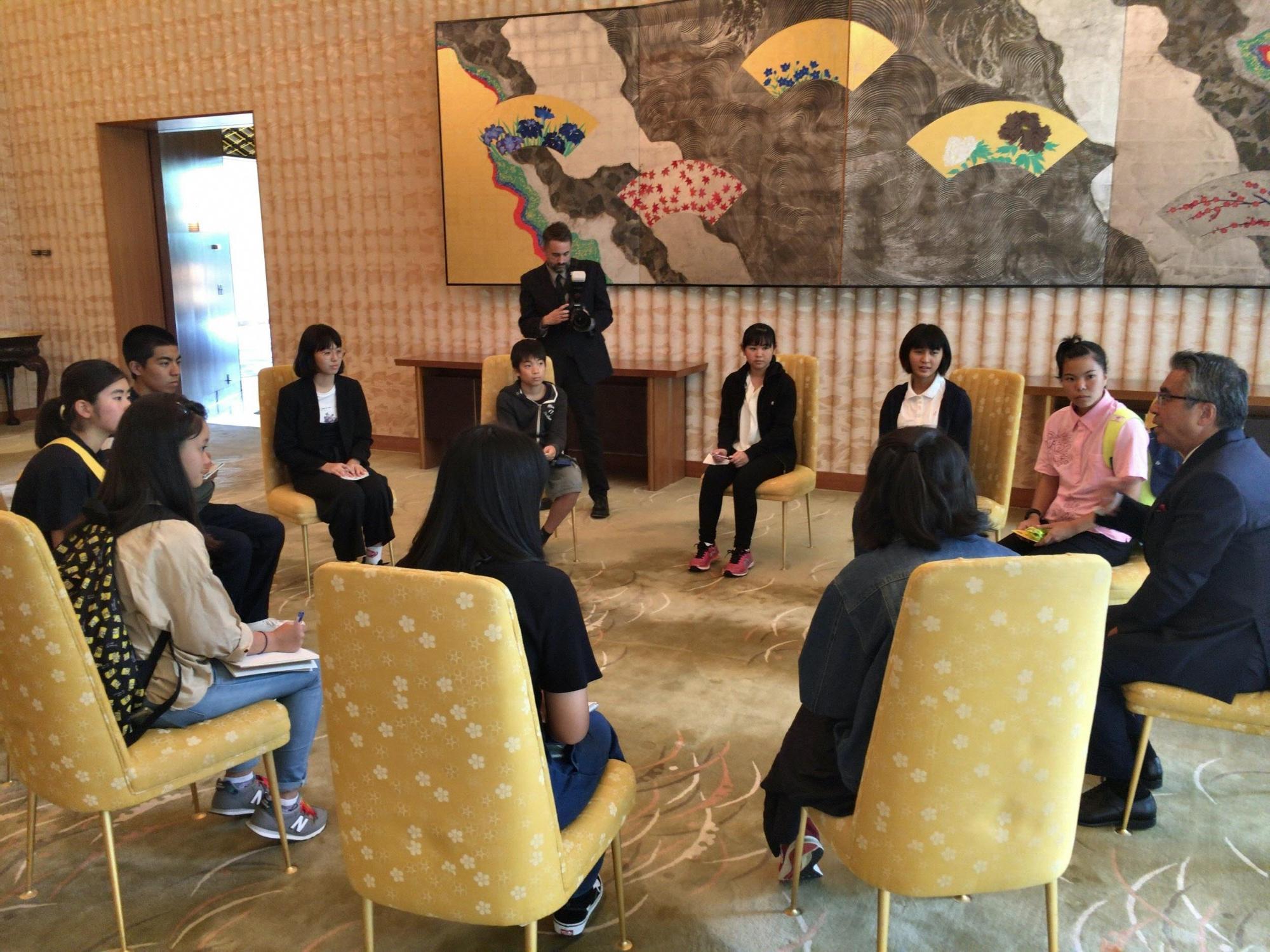 広間のような場所で、黄色の背もたれの椅子に各人が座り在米日本国大使の話を聞いている写真