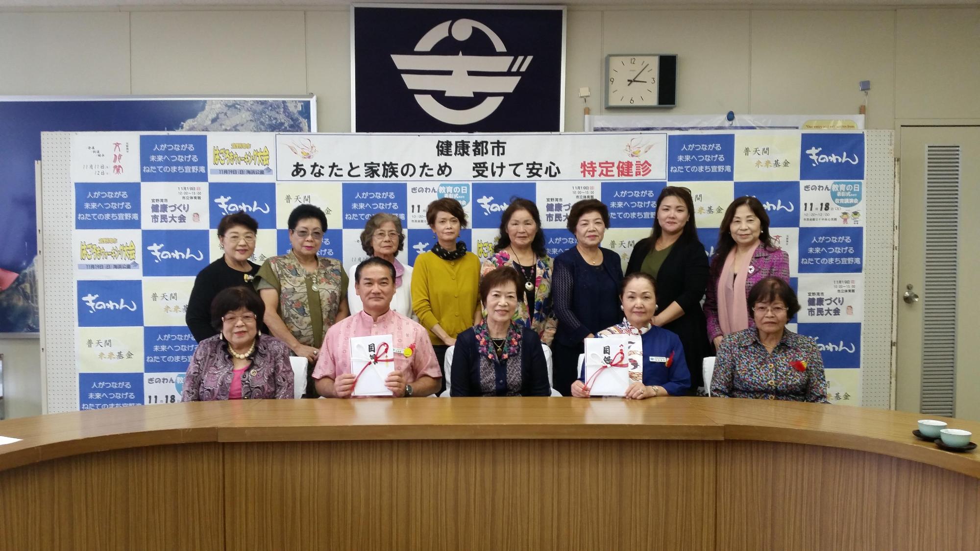 前列に5名の方々が座り、うち佐喜眞市長と青い服を着た女性が目録を持っており、後方には8名の関係者の方々が笑顔で写っている集合写真