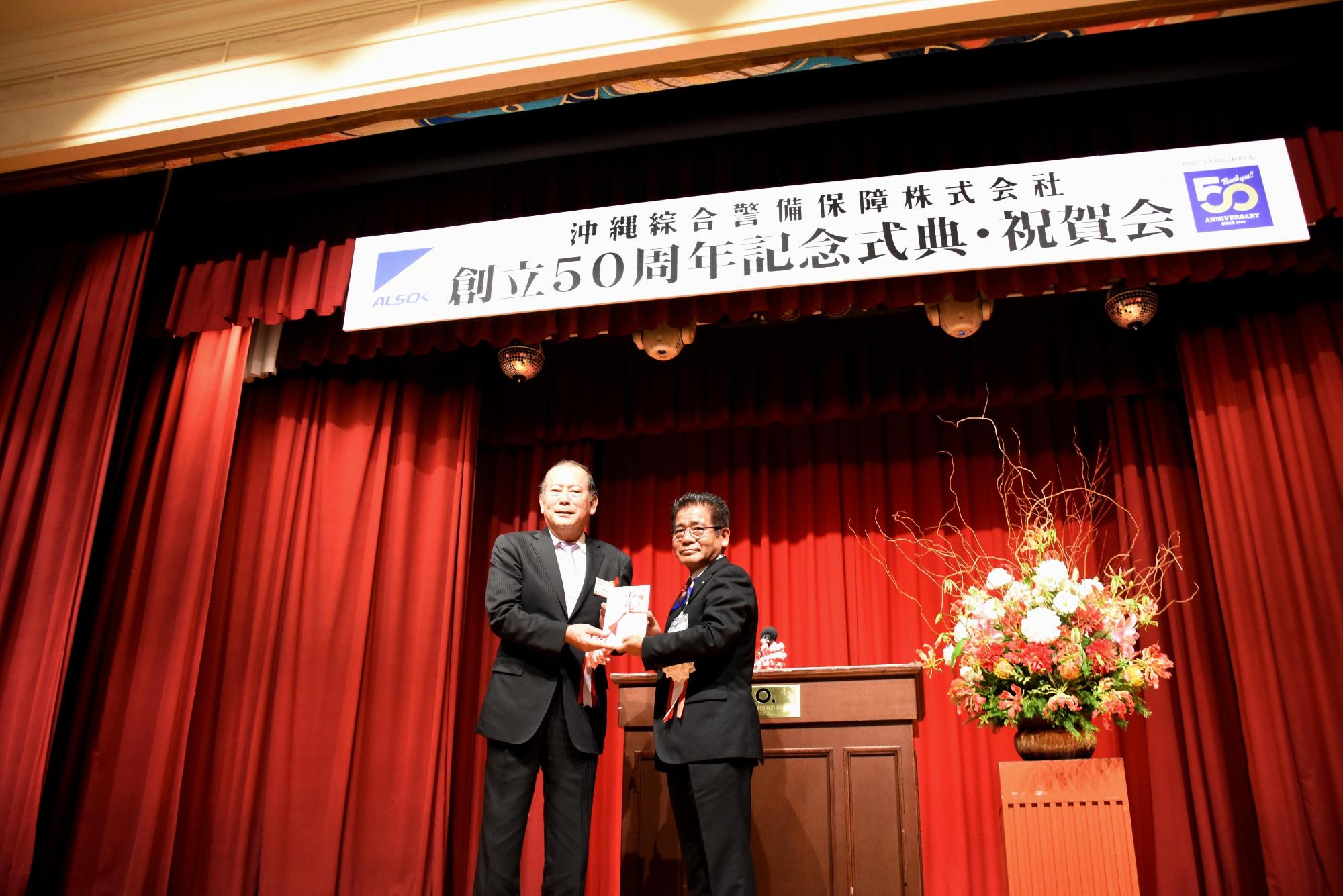 赤色のステージカーテンの舞台上に池田 典夫さんと松川市長が寄附金を持ち、写っている写真