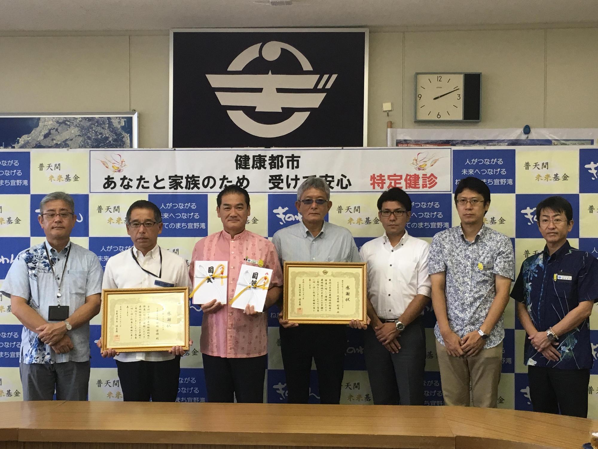 佐喜眞市長が両手に目録を持ち、両隣の男性が額縁に入った賞状を持っており、関係者の方々が両手を前に組んで写っている集合写真