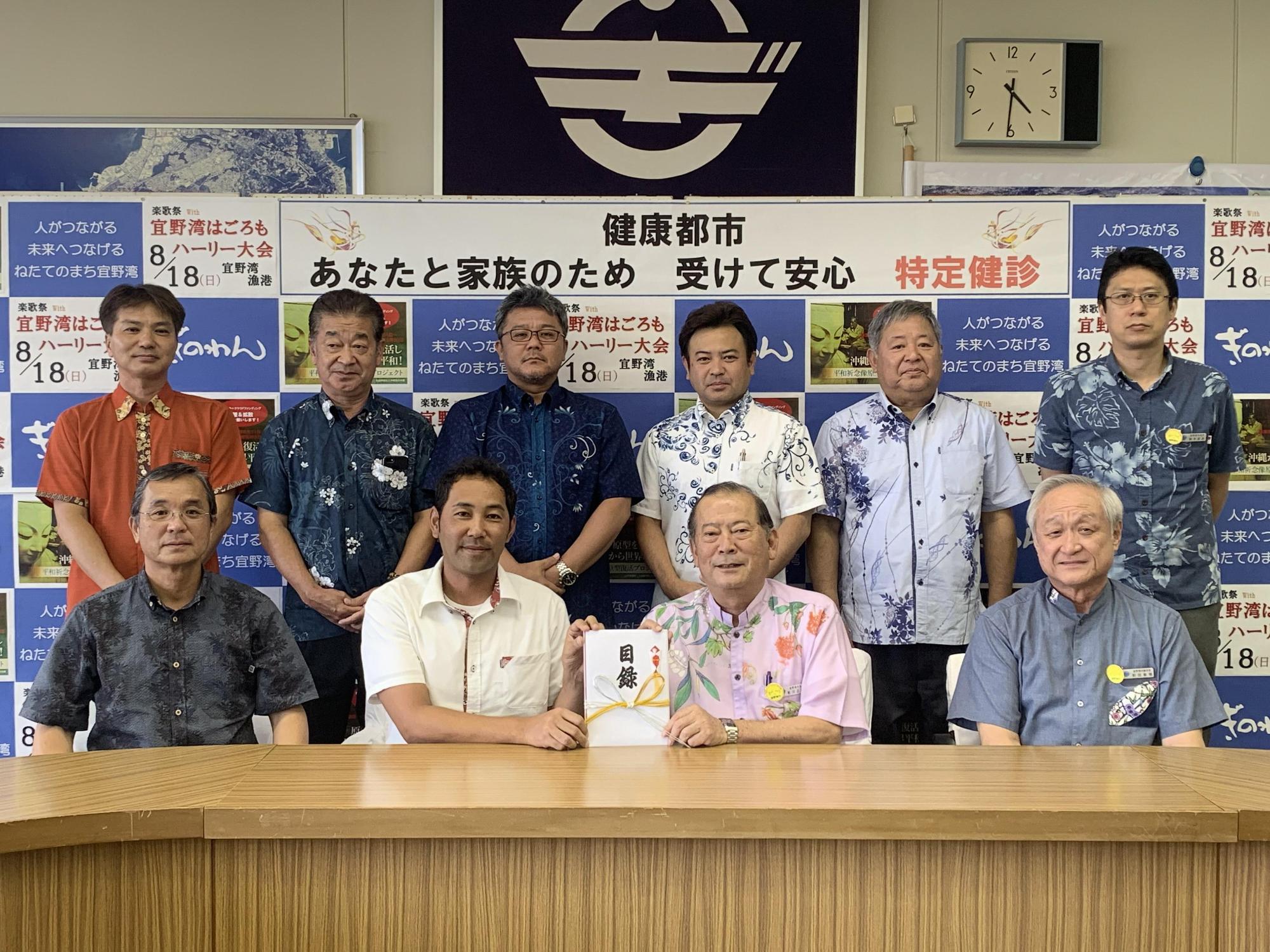 前列に4名の方々が座り、真ん中に座っている松川市長と1名の男性が目録を持っており、後方には6名の関係者の方々が写っている集合写真