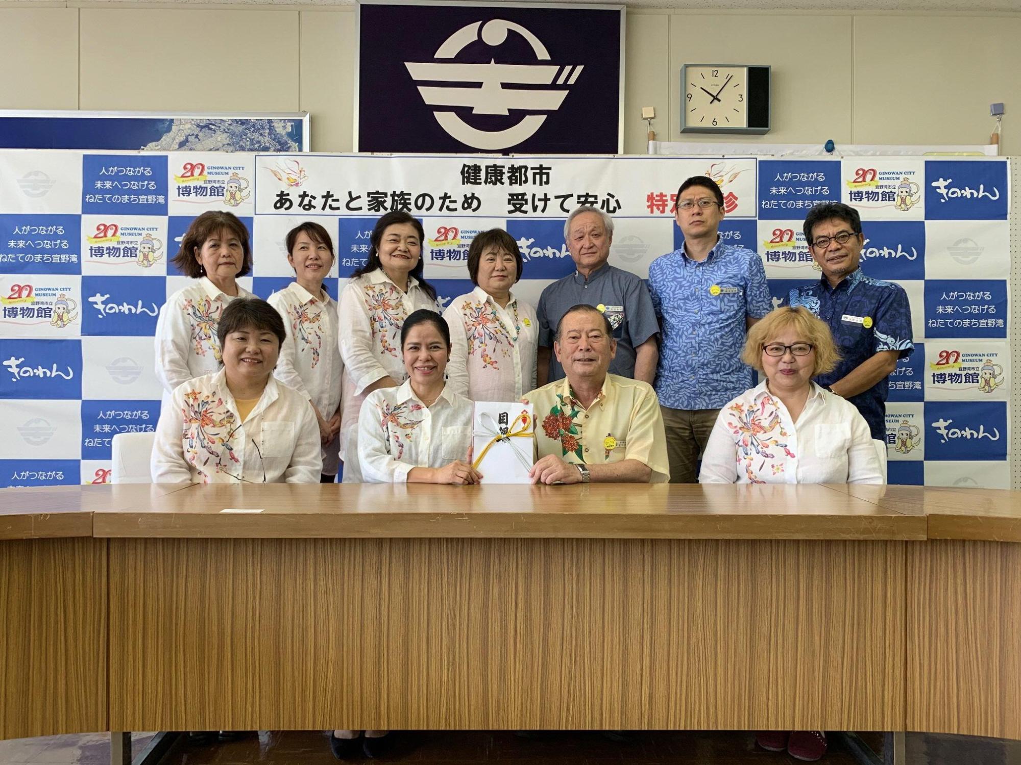 前列に4名の方々が座り、うち松川市長と1名の女性が目録を持っており、後方には関係者の方々が笑顔で写っている集合写真